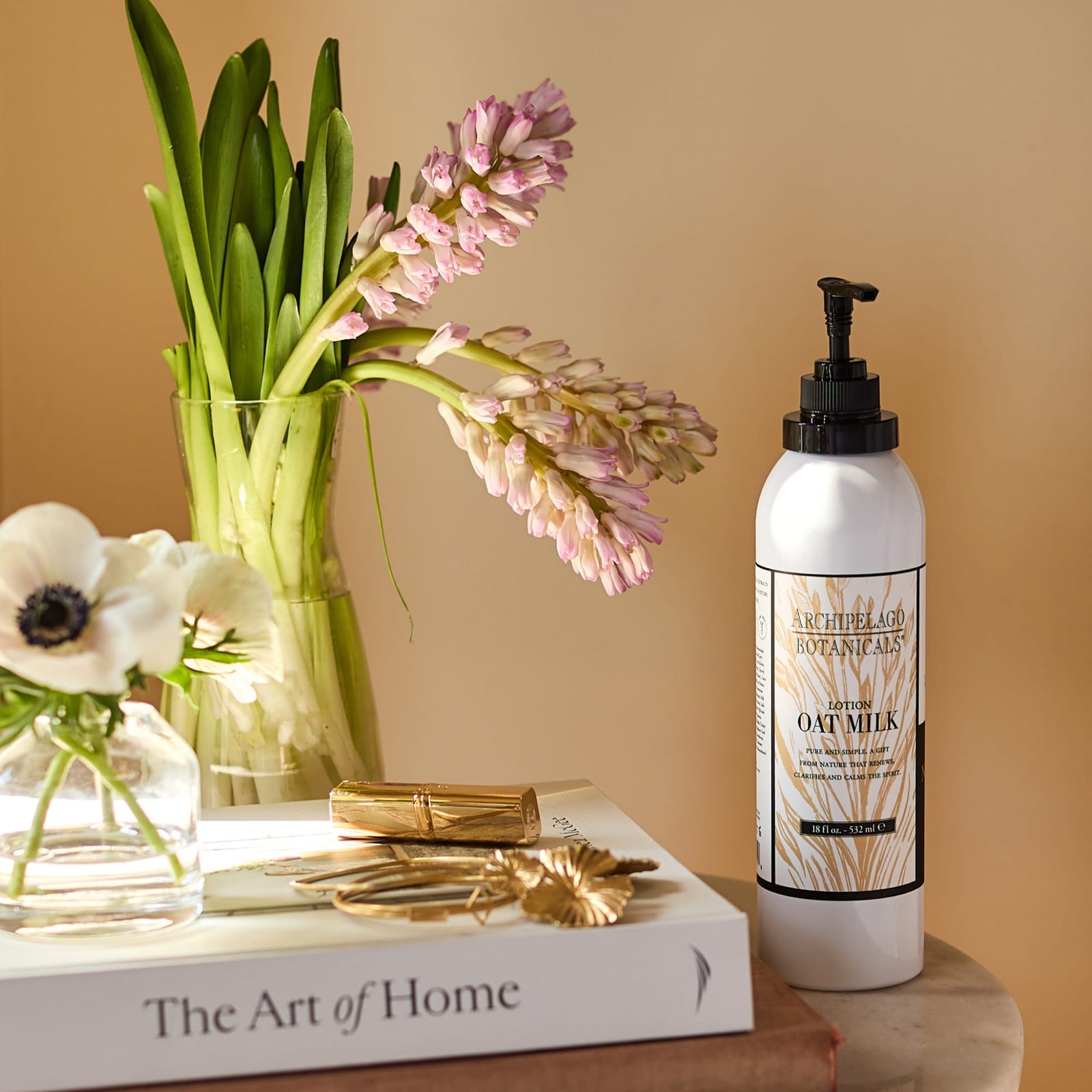 A bottle of Oat Milk Body Lotion rests atop a stack of books next to a vase filled with pink hyacinths and white flowers on a sunlit table. - image 6