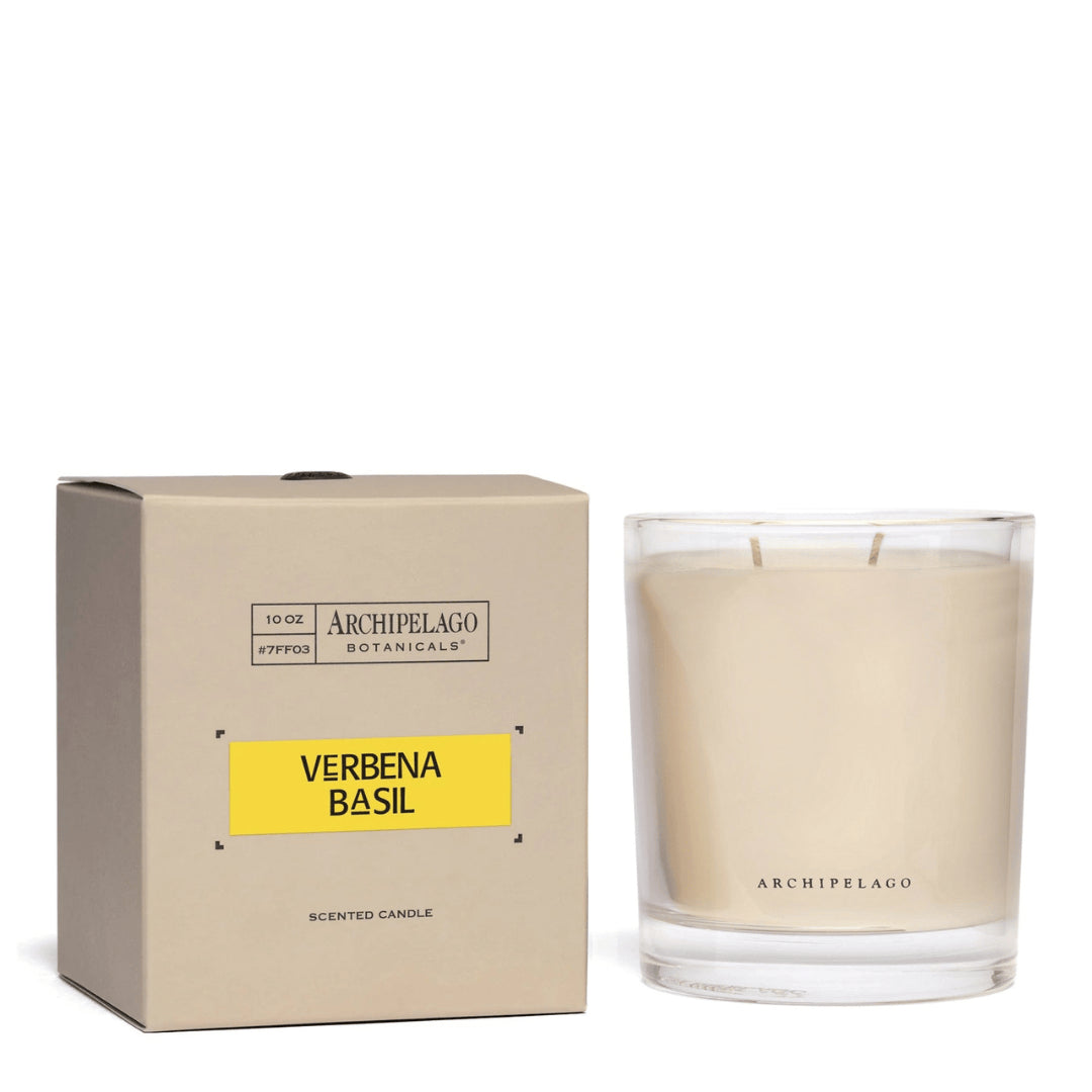 A beige Archipelago Botanicals box labeled Verbena Basil sits next to a clear glass Verbena Basil Credenza Candle filled with white wax. Both items feature the Archipelago Botanicals brand. - image 1