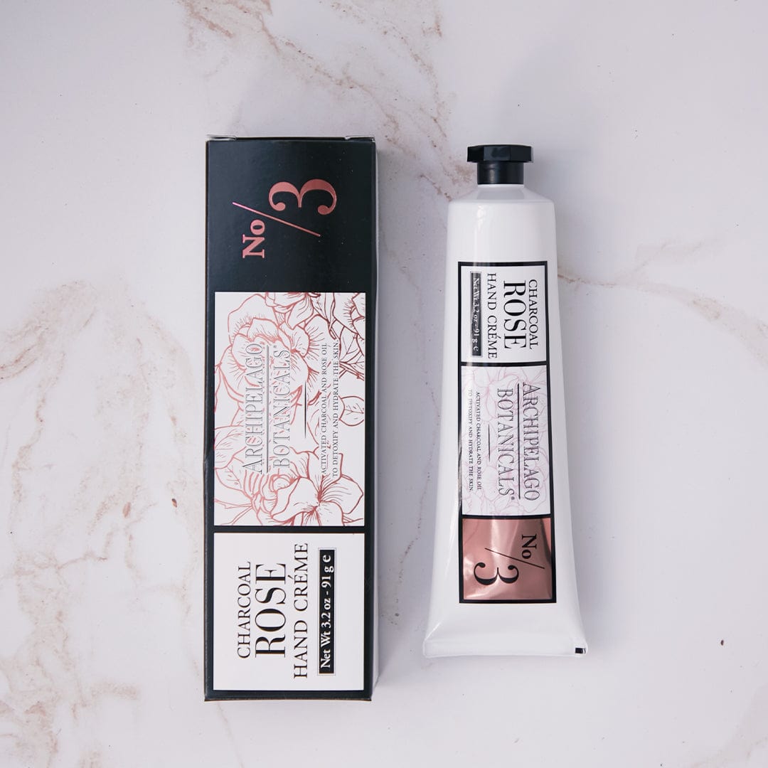 A tube and box of Charcoal Rose Hand Creme are displayed on a marble surface. This hydrating hand cream comes in packaging adorned with floral designs and accented in black, white, and rose gold—perfect for elevating your moisturizing routine. - image 2