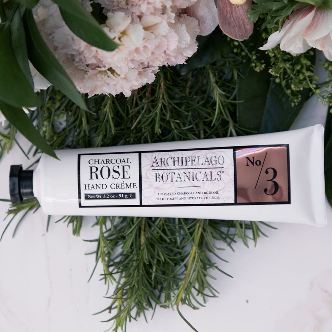 A tube of Charcoal Rose Hand Creme, a hydrating hand cream perfect for any moisturizing routine, lies on green foliage and light pink flowers, with a white and rose gold label reading “No. 3.”. - image 3