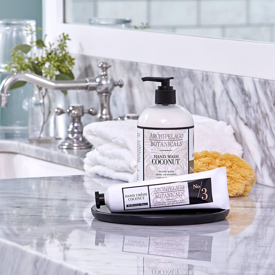Bottles of Coconut Hand Creme by Archipelago Botanicals rest on a marble bathroom counter alongside a white towel, sponge, and small green plant near the sink. This nourishing hand cream hydrates your hands with coconut oil, leaving them soft and refreshed. - image 2