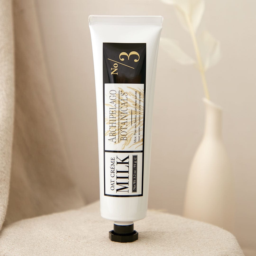 A white tube labeled Oat Milk Hand Crème stands upright on a beige fabric surface, offering moisturizing skincare for dry hands; in the background, a blurred beige vase and plant add a soft touch. - image 2