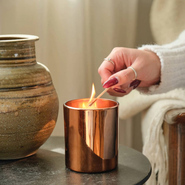 A hand with red nails and rings lights the Pumpkin Macchiato Boxed Candle in a shiny copper holder beside a ceramic vase on a dark table, filling the cozy background with a warm fall fragrance.