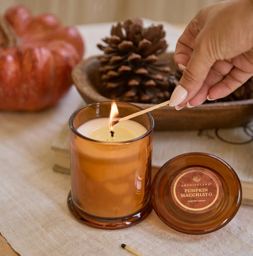 A hand lights the Pumpkin Macchiato Jar Candle with a match, while pinecones and a pumpkin create a softly blurred backdrop. The candle’s lid sits beside the amber glass jar, making this cozy fall candle an ideal accent for your seasonal décor.