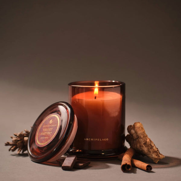 A lit brown glass jar candle labeled “Velvet Harvest Jar Candle” sits with its lid nearby, surrounded by cinnamon sticks, chocolate pieces, a pinecone, and a root on a neutral background—the perfect setting to showcase rich fall fragrances and evoke a cozy seasonal scent.