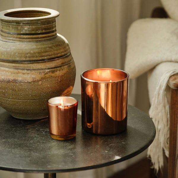 A Velvet Harvest Boxed Votive Candle glows warmly atop a round black side table beside a ceramic vase and a beige textured blanket draped over a wooden chair, filling the space with an inviting fall fragrance.
