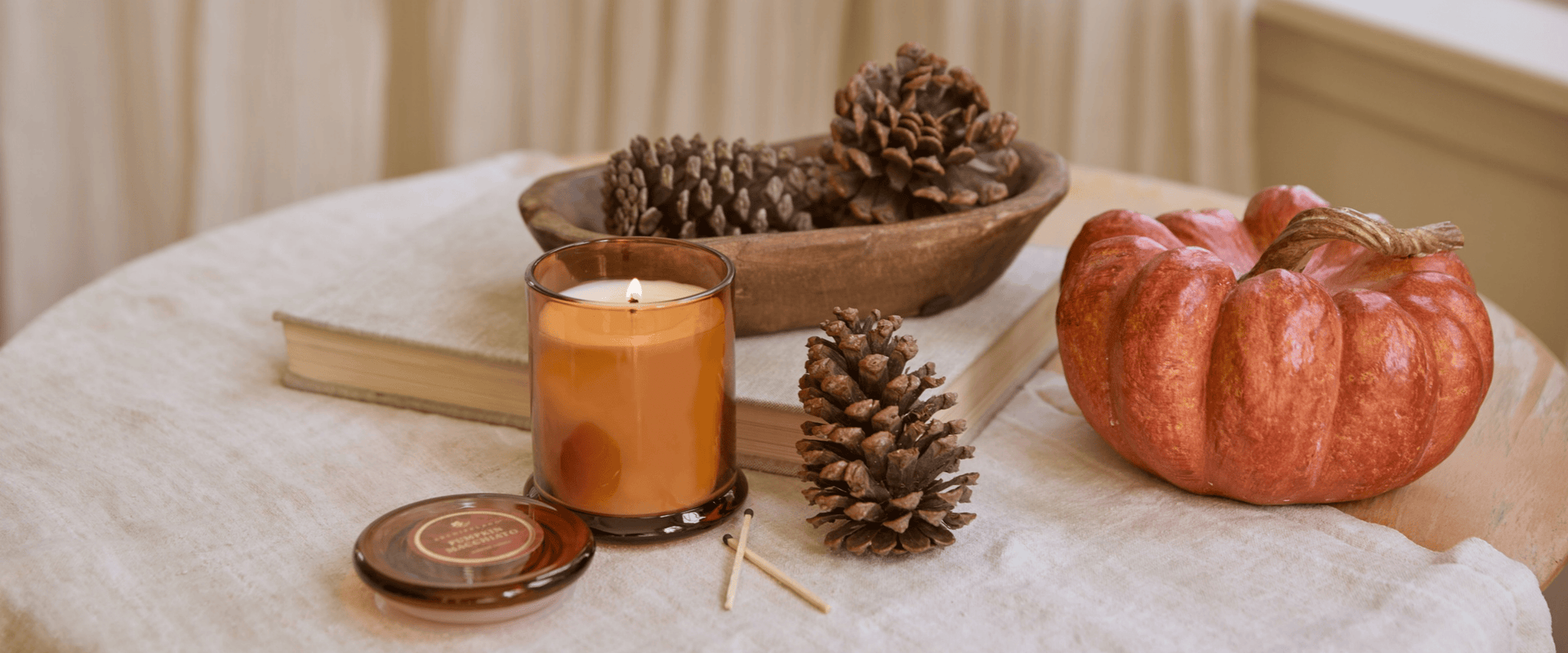 An amber glass jar candle with a lit flame sits on a draped cream fabric surface, surrounded by autumn props including a small orange pumpkin, pinecones, a wooden bowl, books, and a small round container.