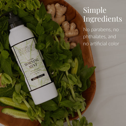 A bottle of Morning Mint Body Lotion sits on a wooden tray alongside fresh mint, cucumber, and ginger. Text reads: Simple Ingredients. Infused with essential oils for aromatherapy benefits. Free from parabens, phthalates, and artificial color.