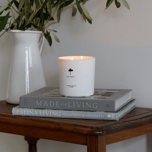 A lit white candle sits atop two stacked books on a wooden table. The top book is Made for Living and the bottom one is Patina Living. There is a white vase with green leaves in the background.