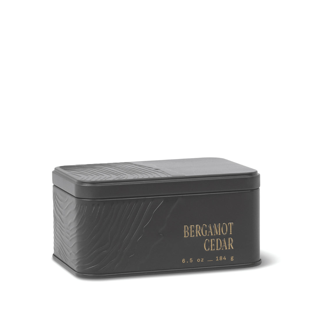 A rectangular black metal travel tin candle with an embossed texture and gold lettering displaying “BERGAMOT CEDAR 6.5 oz – 184 g” on the side. The closed lid keeps the fresh, herbaceous scent of the Bergamot Cedar Travel Tin Candle inside. - image 1