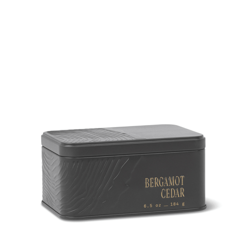 A rectangular black metal travel tin candle with an embossed texture and gold lettering displaying “BERGAMOT CEDAR 6.5 oz – 184 g” on the side. The closed lid keeps the fresh, herbaceous scent of the Bergamot Cedar Travel Tin Candle inside.