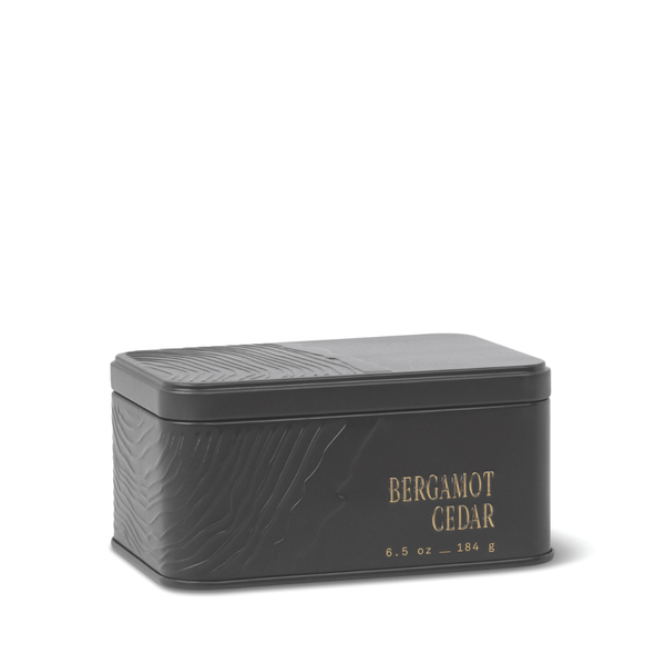 A rectangular black metal travel tin candle with an embossed texture and gold lettering displaying “BERGAMOT CEDAR 6.5 oz – 184 g” on the side. The closed lid keeps the fresh, herbaceous scent of the Bergamot Cedar Travel Tin Candle inside.