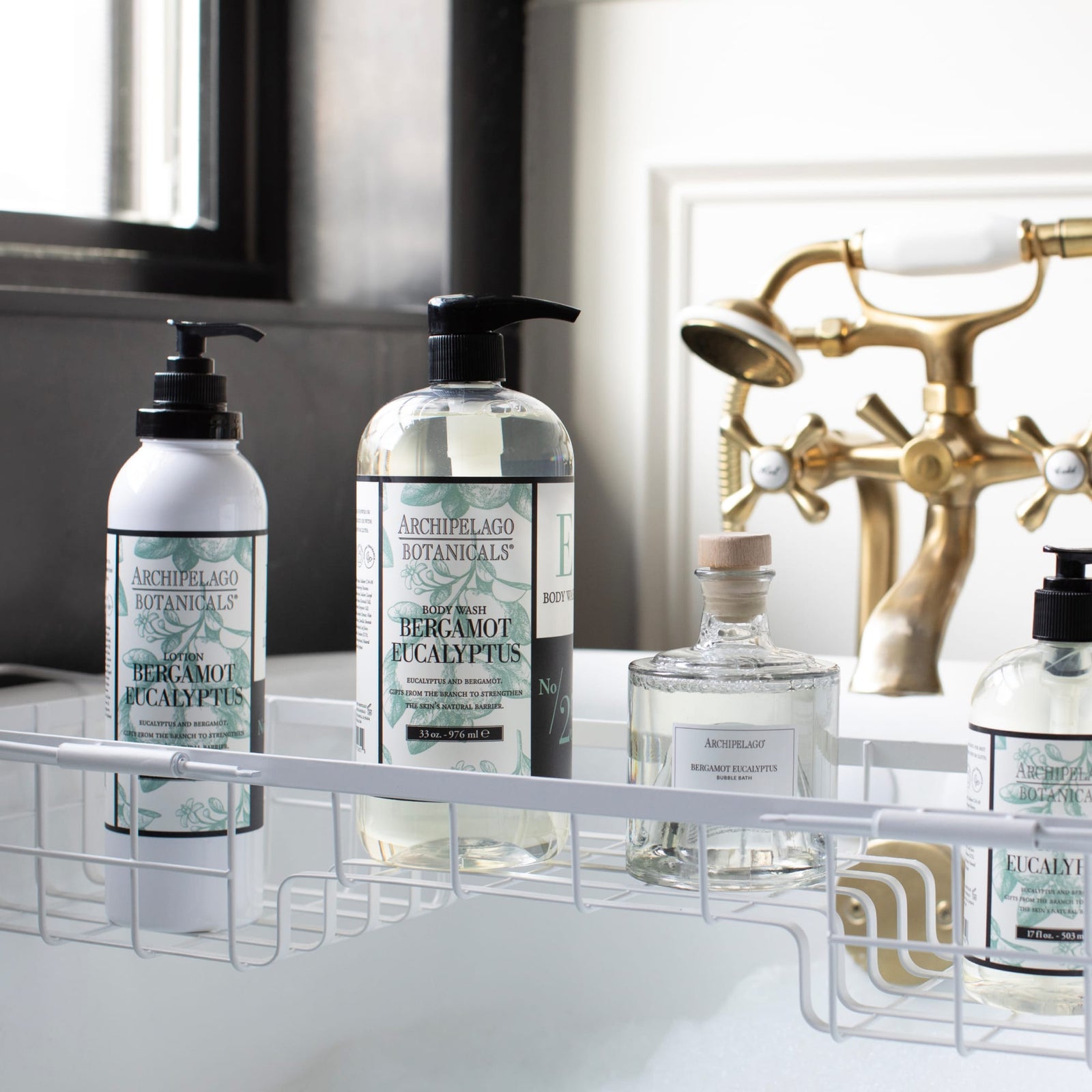 Bottles of Bergamot Eucalyptus Body Lotion are arranged on a white wire rack over a bathtub. A brass faucet and white bathtub are visible in the background near a window.7