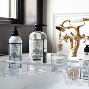 Bottles of Bergamot Eucalyptus Body Lotion are arranged on a white wire rack over a bathtub. A brass faucet and white bathtub are visible in the background near a window.7
