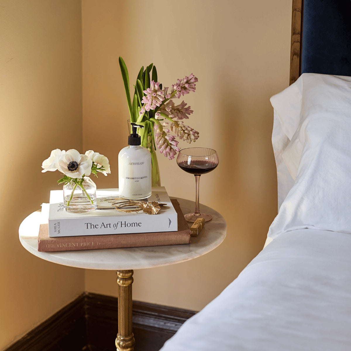 Glass lotion bottle on nightstand with books, glass and flowers.