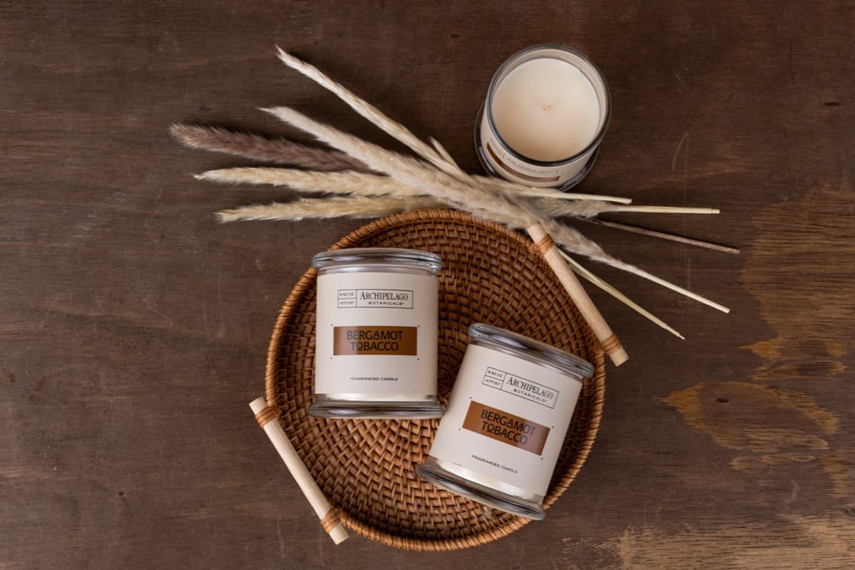 Two Bergamot Tobacco Jar Candles in glass jars sit on a woven tray with dried grass decor, alongside a third soy wax candle above, all arranged on a wooden surface.2