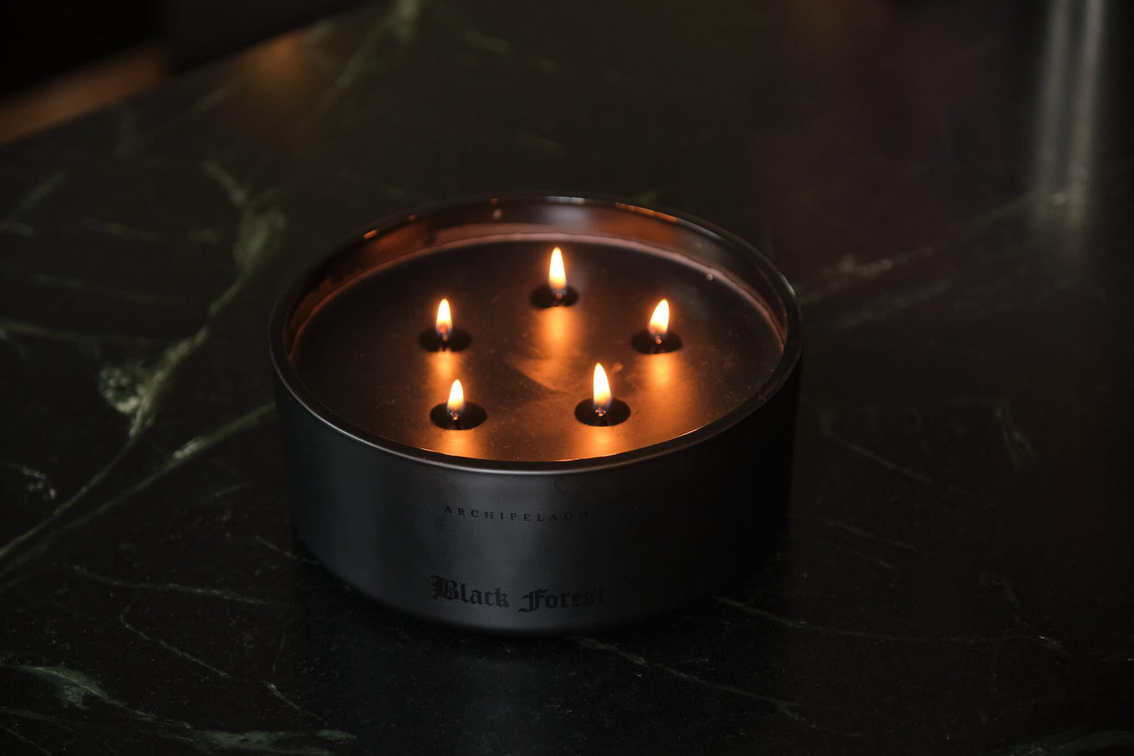 A round, black Forest 5-Wick Candle with five lit wicks, sitting on a dark surface. - image 2