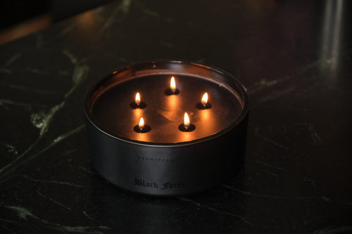 A round, black Forest 5-Wick Candle with five lit wicks, sitting on a dark surface.