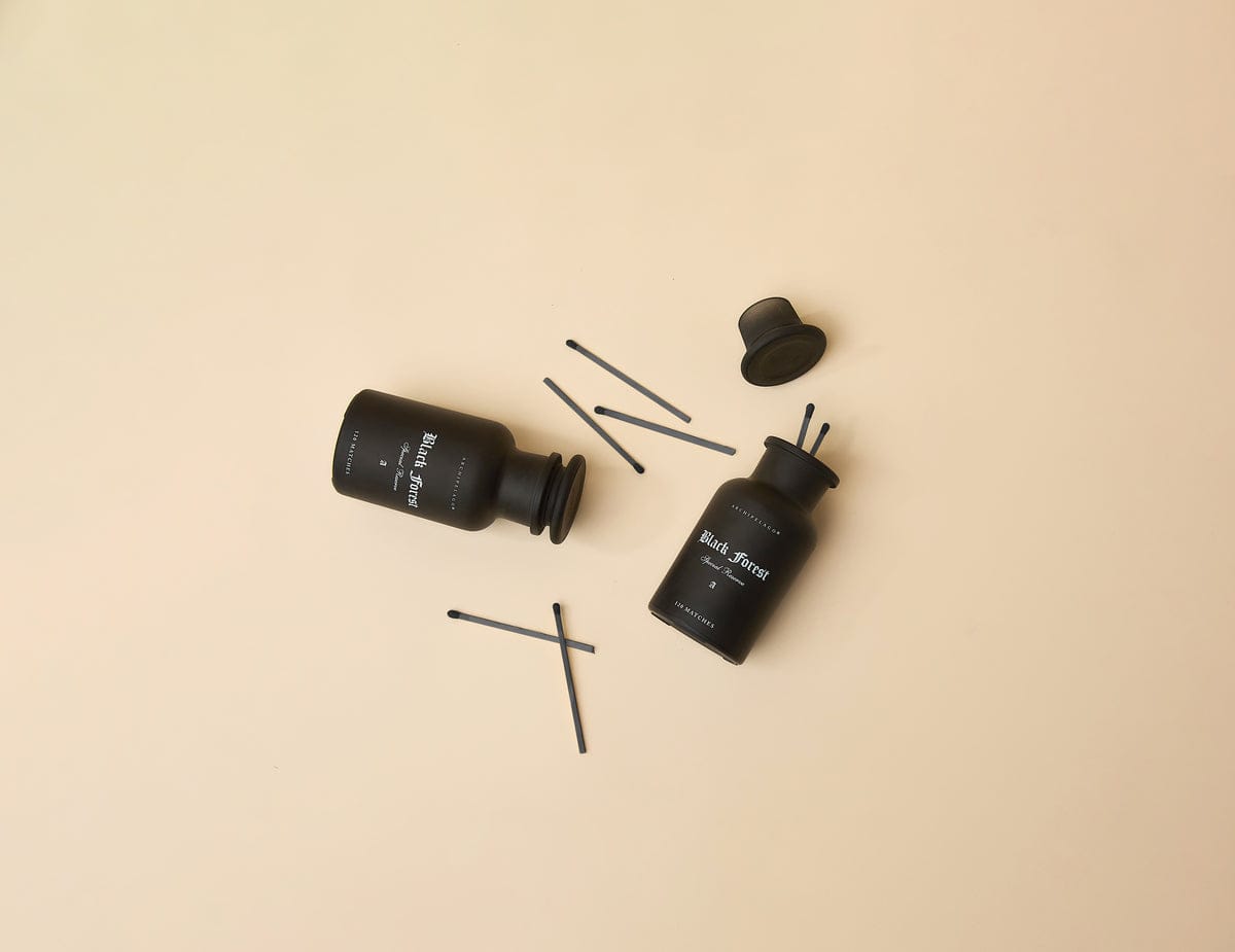 Two Black Forest Luxury Matches apothecary jars—one upright, one tipped over—sit on a beige background with premium matches scattered around. Each matte black jar has a label and a black lid, adding stylish décor to any space.3