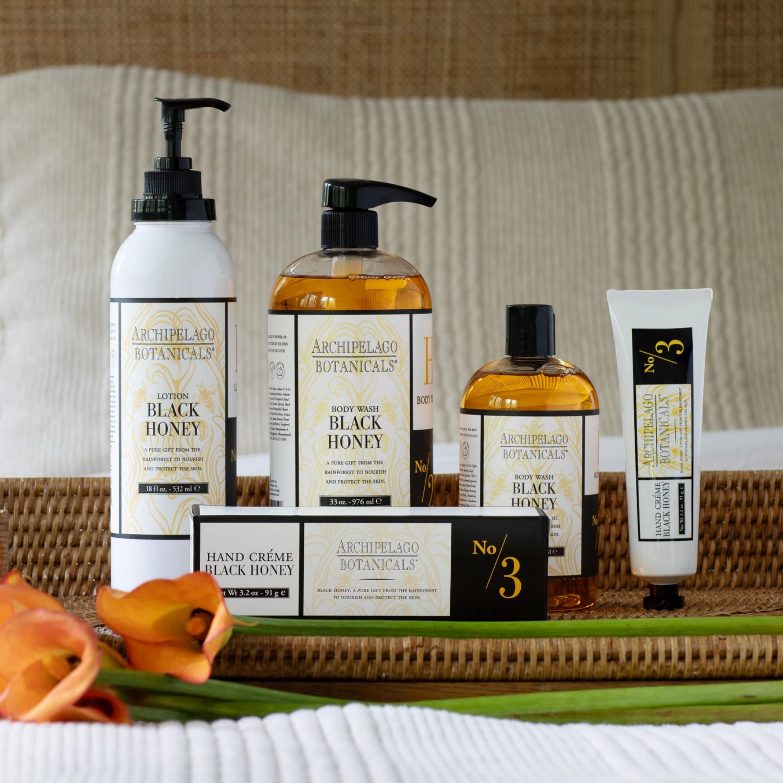 Five Archipelago Botanicals Black Honey products are arranged on a wicker tray atop a bed, with two orange calla lilies nearby. The items include Black Honey Body Lotion, body wash, hand crème, and a small bottle.7