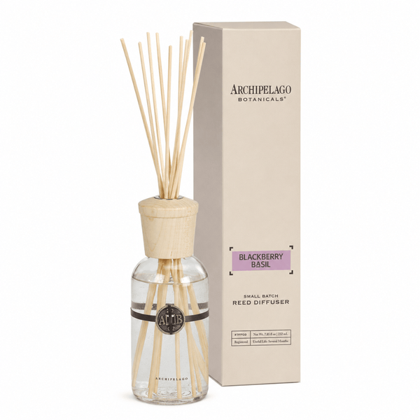 The Blackberry Basil Reed Diffuser with wooden sticks and cap stands next to its beige box, featuring a black logo on the glass bottle filled with a refreshing blackberry basil fragrance blend.