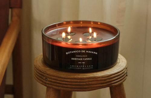 A Botanico de Havana 5-Wick Candle in a low, wide amber glass jar sits on a small wooden stool.