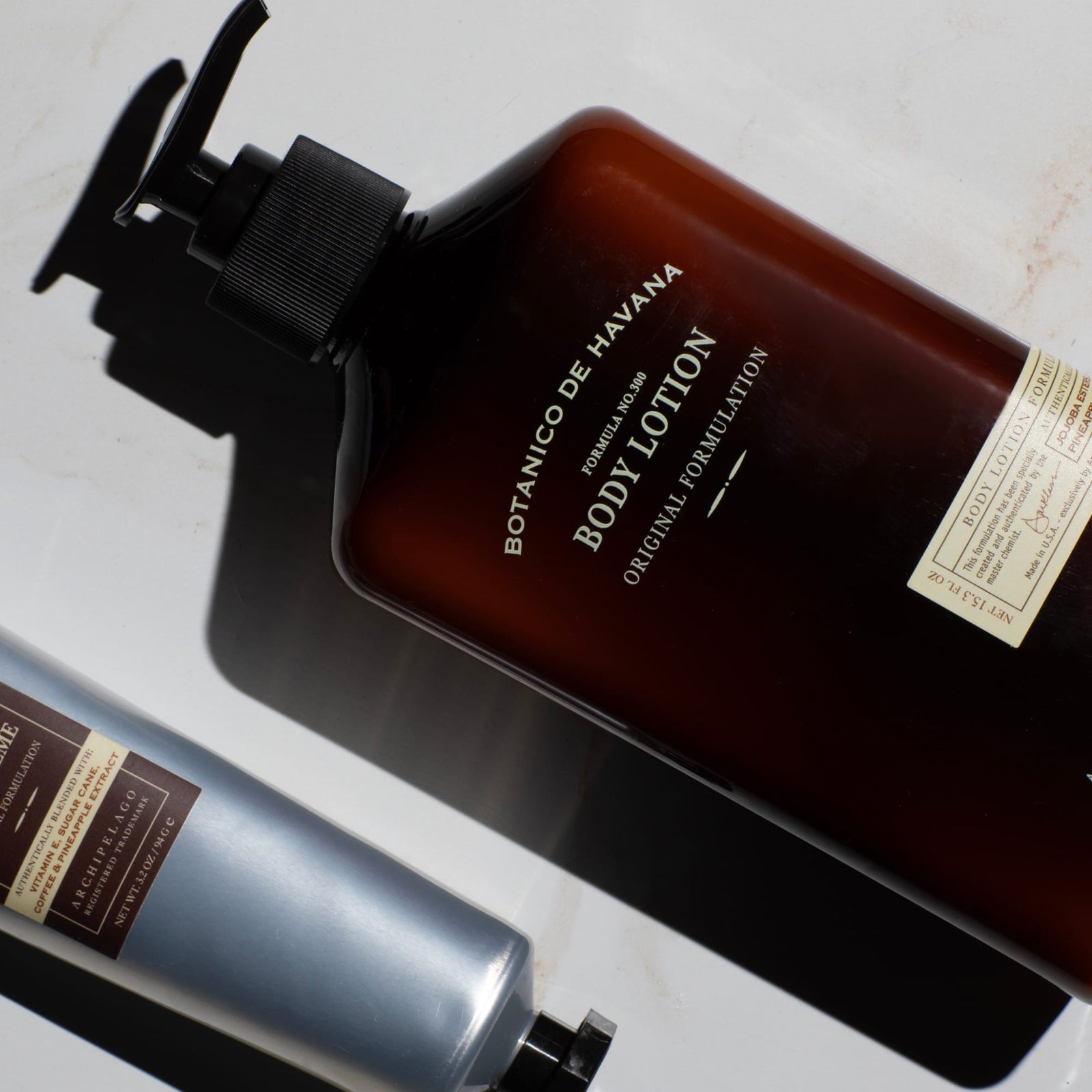 Two bottles of personal care products, including one brown-labeled Botanico de Havana Body Lotion featuring natural beauty ingredients that moisturize dry skin and one silver bottle, are arranged on a white surface with dramatic lighting and shadows.8