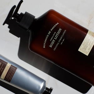 Two bottles of personal care products, including one brown-labeled Botanico de Havana Body Lotion featuring natural beauty ingredients that moisturize dry skin and one silver bottle, are arranged on a white surface with dramatic lighting and shadows.8