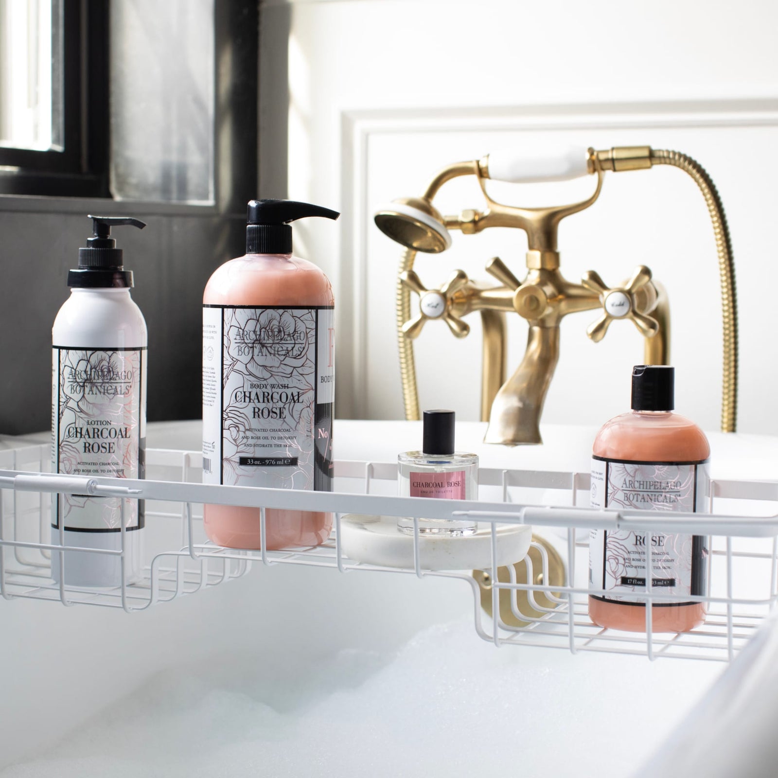 Four charcoal and rose bath products, including a luxurious Charcoal Rose Body Lotion, are arranged on a white metal bath tray over a bubble-filled tub, with gold faucet fixtures and natural light in the background.7