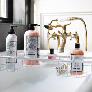 Four charcoal and rose bath products, including a luxurious Charcoal Rose Body Lotion, are arranged on a white metal bath tray over a bubble-filled tub, with gold faucet fixtures and natural light in the background.7