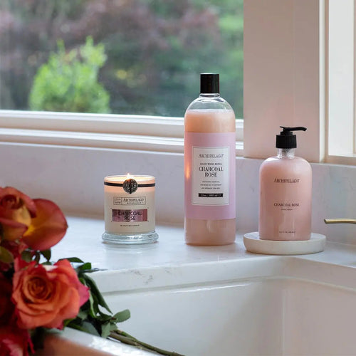 A lit candle, a bottle of Charcoal Rose Hand Wash Refill, and eco-friendly hand wash rest on a bathroom counter by a window, accented with a bouquet of orange roses.