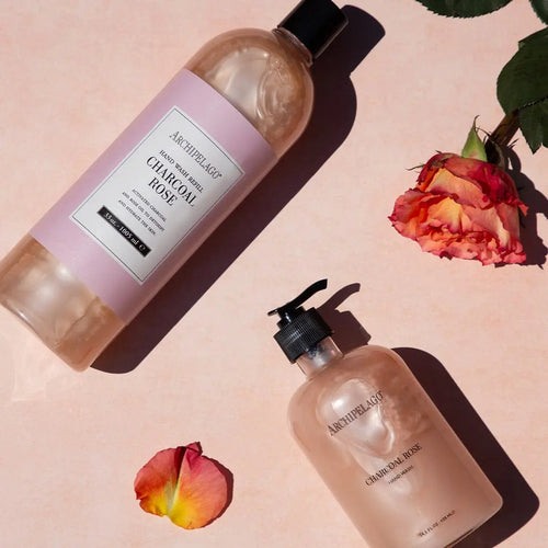 A Charcoal Rose Hand Wash Refill and hand wash with activated charcoal rest on a pink surface. A rose, rosebud, and petals nearby enhance the eco-friendly vibe.