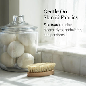 A glass jar of Charcoal Rose Laundry Detergent sits on a white countertop next to a wooden brush. Text reads: Gentle on skin & fabrics, with plant-derived enzymes. Free from chlorine, bleach, dyes, phthalates, and parabens.5