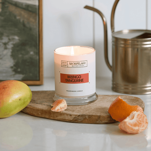 A lit Mango Tangerine scented candle sits on a wooden board next to a fresh mango, peeled tangerine segments, and an orange peel, with a metal watering can in the background.
