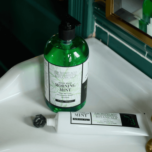 A bottle of Archipelago Botanicals Morning Mint hand wash and a matching mint hand cream tube sit on a white sink beside a green-tiled wall and a mirror.