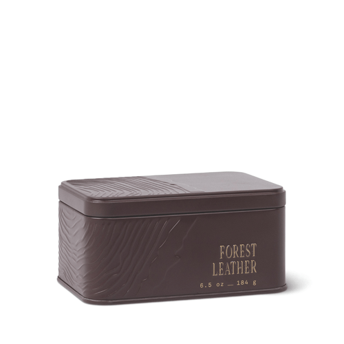 A rectangular, dark brown Forest Leather Travel Tin Candle featuring embossed patterns and gold text on the front that reads “Forest Leather candle 6.5 oz – 184 g.” The lid and sides are adorned with a textured design, reflecting the candle’s rich sandalwood notes.