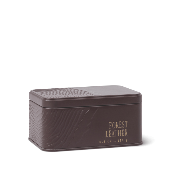 A rectangular, dark brown Forest Leather Travel Tin Candle featuring embossed patterns and gold text on the front that reads “Forest Leather candle 6.5 oz – 184 g.” The lid and sides are adorned with a textured design, reflecting the candle’s rich sandalwood notes.