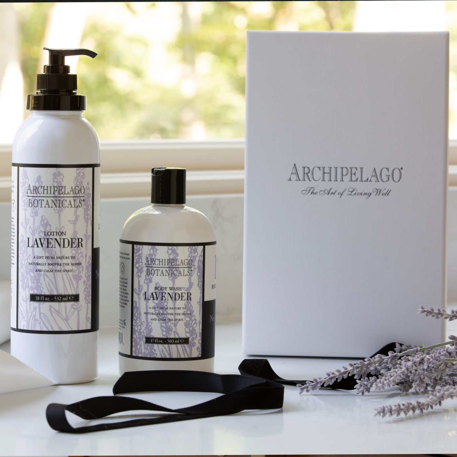 Lavender Body Lotion bottles from Archipelago Botanicals, enriched with essential oils, are displayed next to a white Archipelago gift box, black ribbon, and sprigs of lavender against a soft outdoor background. This moisturizing lotion brings a touch of luxury to any self-care routine.7
