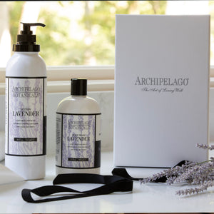 Lavender Body Lotion bottles from Archipelago Botanicals, enriched with essential oils, are displayed next to a white Archipelago gift box, black ribbon, and sprigs of lavender against a soft outdoor background. This moisturizing lotion brings a touch of luxury to any self-care routine.7