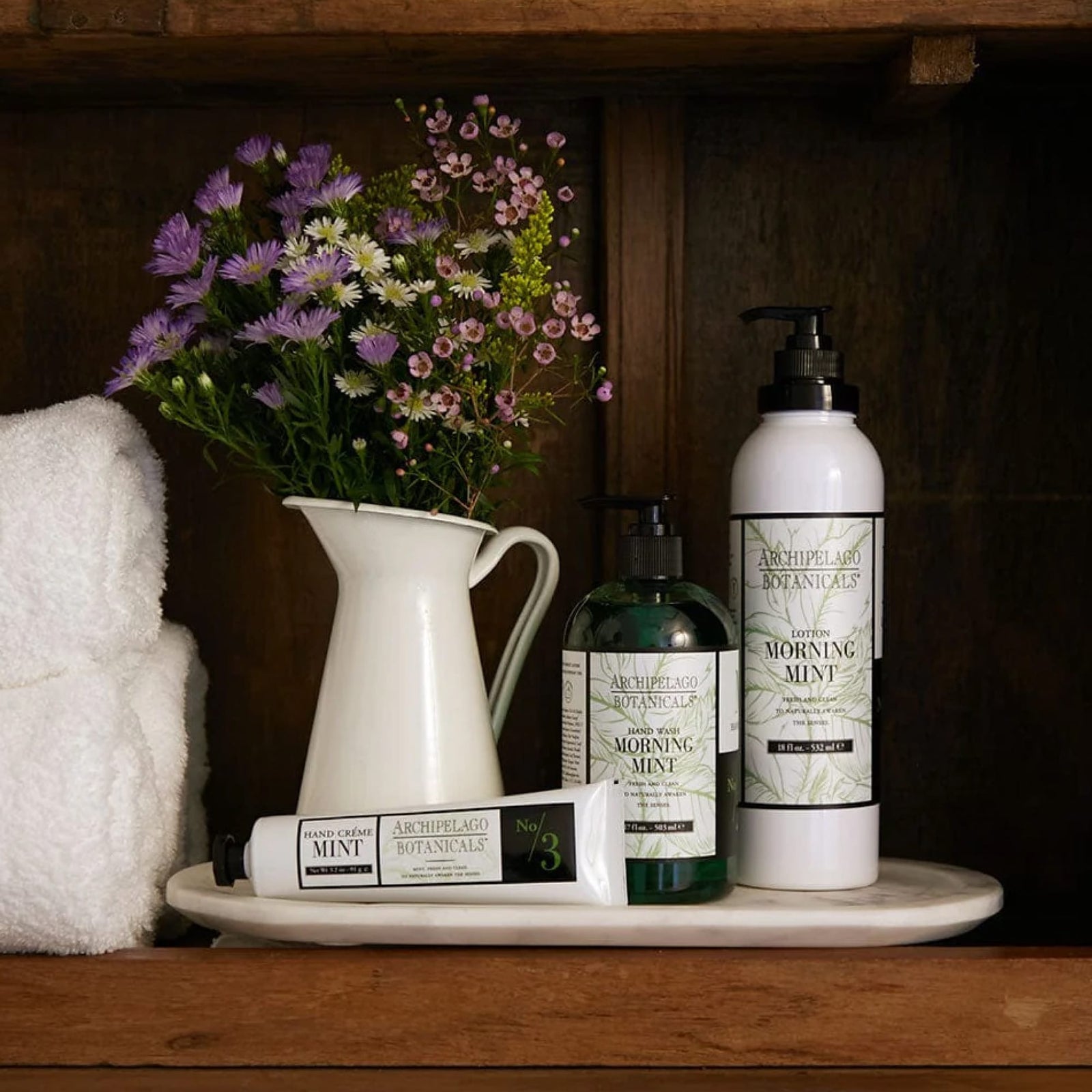 A white pitcher adorned with purple and pink flowers sits beside white towels and a trio of Archipelago Botanicals bath products, including the Morning Mint Body Lotion enriched with essential oils, all arranged on a wooden shelf.7