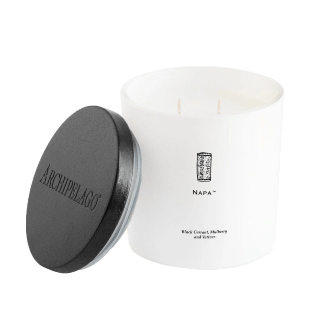 The Napa Luxe Candle is a fruity-scented coconut wax candle in a glass jar labeled NAPA, with notes of black currant, mulberry, and vetiver. It has a black lid reading ARCHIPELAGO, shown leaning against the white jar.1