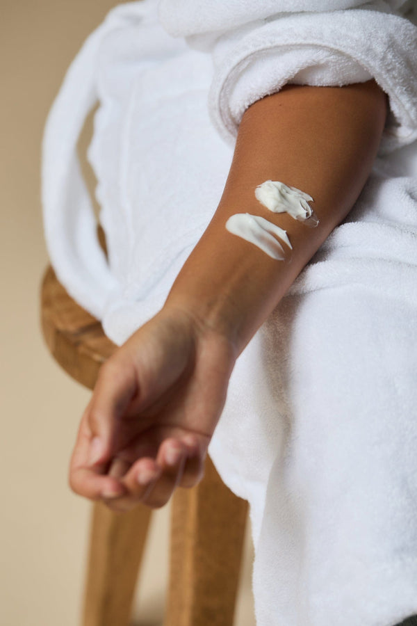 Person applying Body Butter to their arm with a white towel draped over them, sitting on a wooden stool.