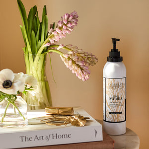 A bottle of Oat Milk Body Lotion rests atop a stack of books next to a vase filled with pink hyacinths and white flowers on a sunlit table.6