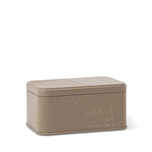 Introducing the Olivewood Ash Travel Tin Candle: a rectangular taupe tin featuring textured lines and “OLIVEWOOD ASH” embossed on the front, along with its weight—6.5 oz (184 g). The smooth lid, finished in a matching taupe hue, beautifully complements the understated mineral fragrance inside. - image 1