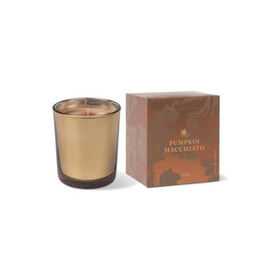 A gold-toned Pumpkin Macchiato Boxed Votive Candle in a glass holder sits next to its brown packaging, which is adorned with subtle floral patterns and elegant gold text. Crafted from coconut wax, this fall candle combines autumnal charm with stylish design. - image 1