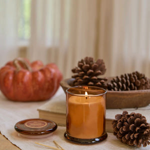 An amber glass jar candle with a lit flame sits on a table runner, styled with a small pumpkin, pinecones, books, and a lid in the foreground. - image 2