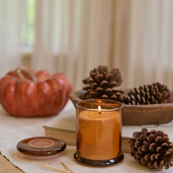 An amber glass jar candle with a lit flame sits on a table runner, styled with a small pumpkin, pinecones, books, and a lid in the foreground.