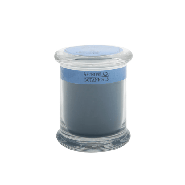 The Santorini Jar Candle features solid blue-gray wax infused with essential oils in a glass apothecary jar. It has a blue label reading Archipelago Botanicals and comes with a matching glass lid.