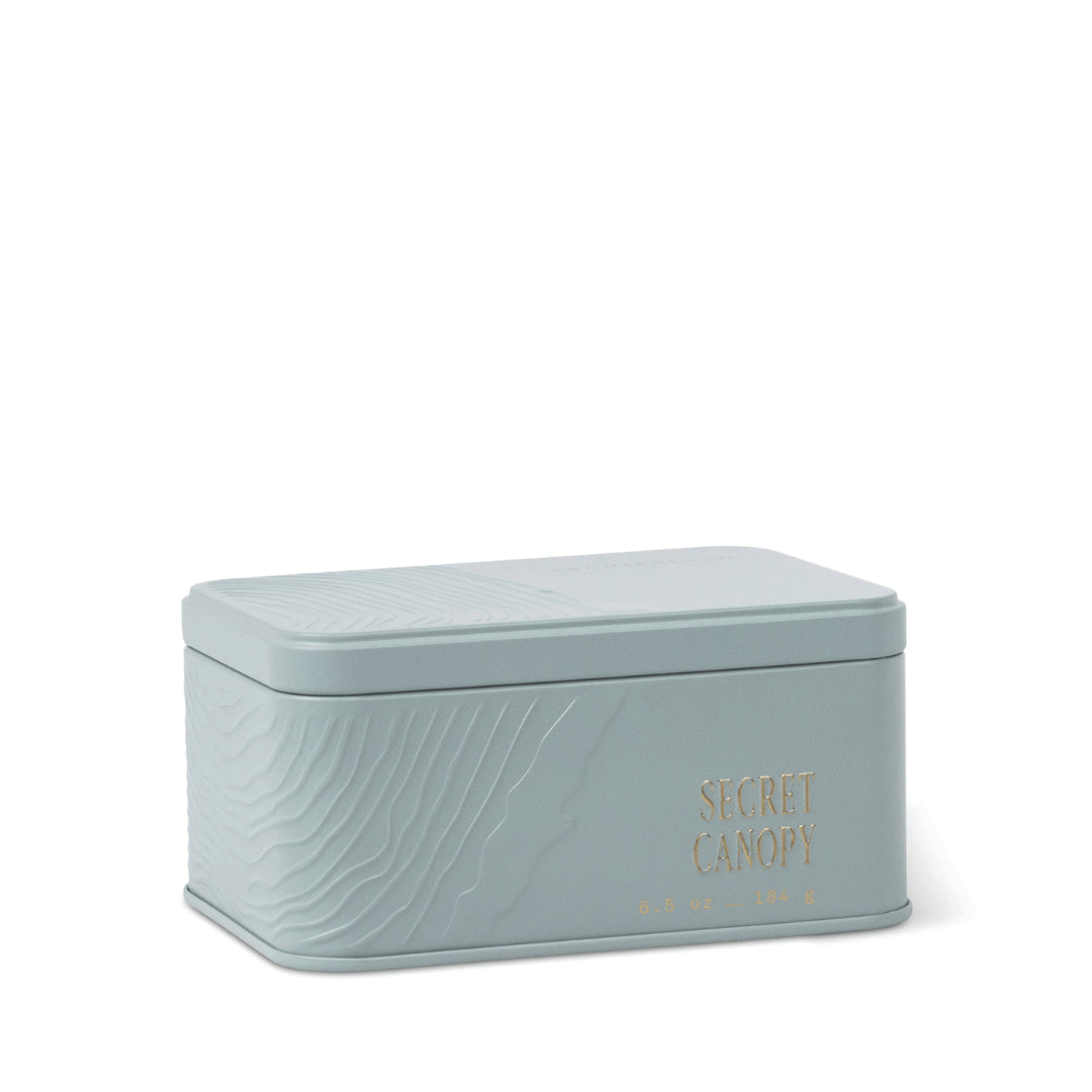 A light gray, rectangular Secret Canopy Travel Tin Candle with embossed wave patterns, gold “SECRET CANOPY” lettering, and a fitted lid rests against a white background. This travel candle fills any space with a refreshing mineral fragrance wherever you journey. - image 1