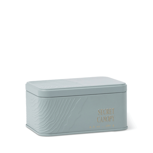 A light gray, rectangular Secret Canopy Travel Tin Candle with embossed wave patterns, gold “SECRET CANOPY” lettering, and a fitted lid rests against a white background. This travel candle fills any space with a refreshing mineral fragrance wherever you journey.